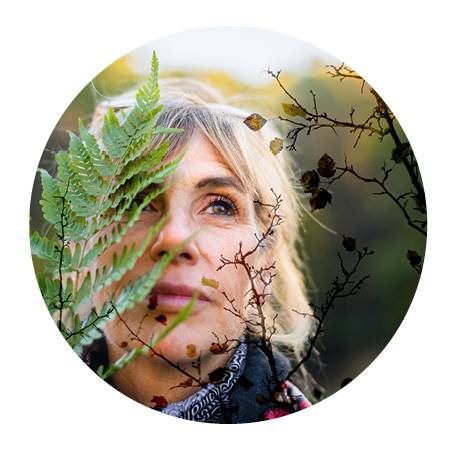 Maria Gambino standing amongst the forest with a green fern covering part of her face looking offset into the distance.